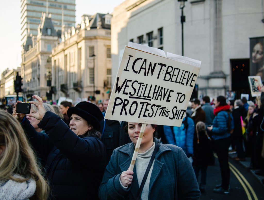 A crowd of demonstrators on te street. A young woman in the forefront has a hand-written placard that reads “I can’t believe we still have to protest this sh*t”