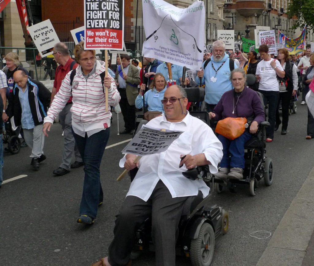 The photo shows a street protest against disability cuts. 20-30 different people holding placards are marching through London, including 3 or 4 powerchair and wheelchair users, a visually impaired person with a white stick and a person with Down Syndrome.
Photo credit: helen.2006 - In Memoriam's photo, licensed under Creative Commons as Attribution-NonCommercial-ShareAlike. Downloaded from Flickr.