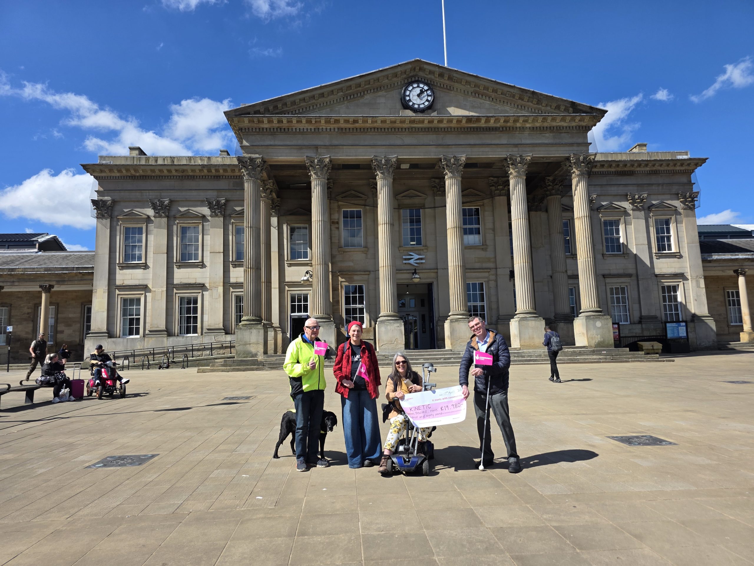 KiNETIC Trustees hold a giant National Lottery Community Fund symbolic cheque in front of Huddersfield train station.